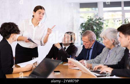 Smart young woman sharing ideas in front of office colleagues of different ages taking notes at table with laptops Stock Photo