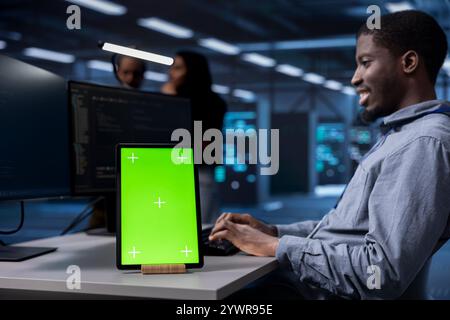 Smiling man working on green screen tablet in modern data center. Happy IT specialist doing IT infrastructure management on server racks in high tech environment using chroma key device Stock Photo