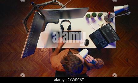Top down view of engineer doing multitasking to finish developing PC code, drinking coffee, listening music. IT admin wearing headphones, solving database errors on computer and laptop, aerial shot Stock Photo
