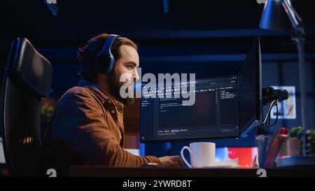Cheerful programmer listening music while doing web development during remote job shift in neon lit home office, finishing coding project. IT expert enjoying audio podcast, working on PC, camera B Stock Photo