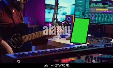 Male artist learning to play guitar on online lesson tutorial, practicing acoustic instrument and looking at phone with greenscreen display. Music producer doing rehearsal guitar play. Camera A. Stock Photo