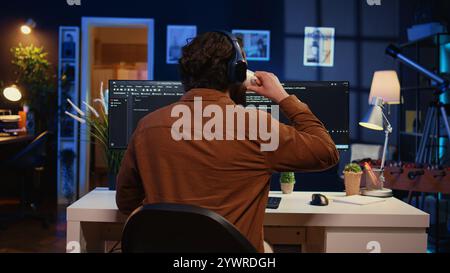 Man doing IT support job from apartment office, drinking cup of coffee and listening music while coding. Developer with headphones on enjoying hot tea and writing code on computer, camera B close up Stock Photo