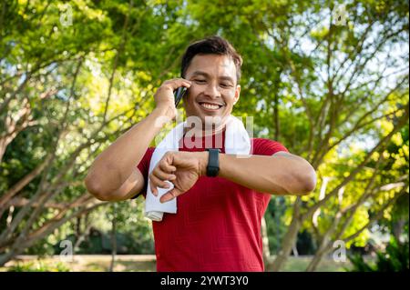 A healthy, happy Asian man in sportswear checks his calorie count on his smartwatch while talking on the phone and jogging in a park on a bright day. Stock Photo