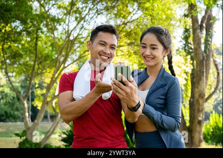 Two happy Asian friends, a female and a male, in sportswear, watch a video together on a smartphone while exercising in a park on a sunny day. people Stock Photo