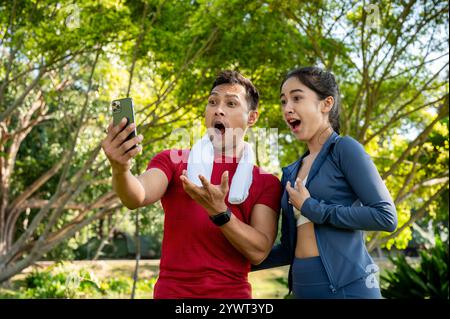 Two shocked, excited Asian friends, a female and a male, in sportswear, watch a video together on a smartphone while exercising in a park on a sunny d Stock Photo