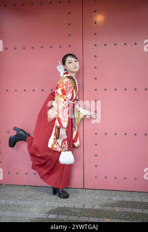 A Japanese woman in her 20s wearing a red kimono (Hakama), a staple of Japanese university graduates, stands in front of a large red iron door and is Stock Photo