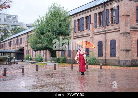 A Japanese woman in her 20s, wearing a red kimono (hakama), a staple of Japanese university graduates, poses in the rain with a red Japanese umbrella Stock Photo