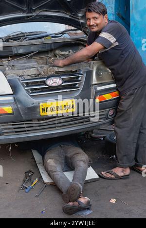 Two young Indian car repair men in Mumbai, India, working on the engine of a car, one of them working underneath the vehicle Stock Photo