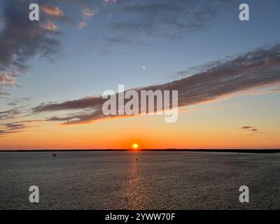 The sun touches the horizon over dark bay water with a narrow golden reflection, below a band of gray cloud, in Erie, Pennsylvania. Stock Photo