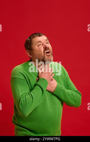 Mature man holding hands on neck feeling sore throat against vibrant red background. Man feels bad, having first symptoms of sick. Stock Photo