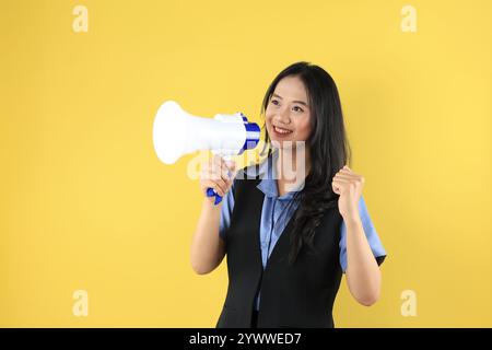 Smiling Young Asian Woman Shouting at Megaphone with Clemched Hand. Concept of Excitement and Young Energy. Isolated on Yellow Background Stock Photo
