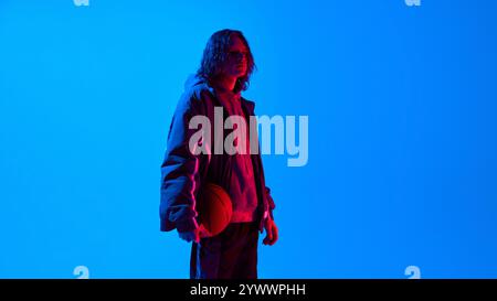 Side view portrait of handsome young man with long curly hair, wearing oversized sportive clothes standing with basketball against blue studio Stock Photo