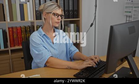 Young woman typing on a computer in an office with bookshelves in the background Stock Photo
