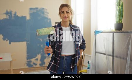 Woman painting living room in new home, using roller brush, wearing casual clothing, surrounded by interior decor in modern apartment, smiling and con Stock Photo