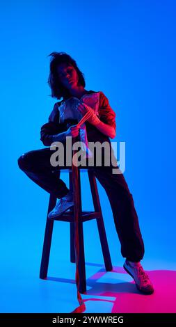 Confident young woman sitting on stool wrapping her hands in boxing tape, dressed in sporty black and white attire against blue studio background in Stock Photo