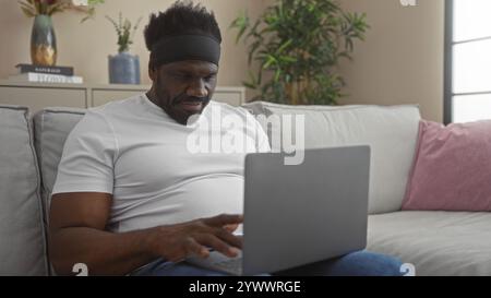 A handsome young african american man with a beard is sitting in the living room of his apartment, working on a laptop while wearing a white t-shirt, Stock Photo