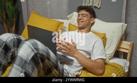 A young, hispanic man lies in bed in a bedroom, smiling while working on a laptop indoors, with cozy pillows and blankets around him. Stock Photo