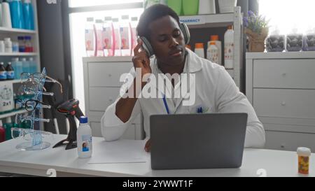 Young man in pharmacy using laptop with headphones in a store environment featuring eyeglasses display and medical products Stock Photo