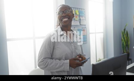 Woman smiling indoors in a bright office setting while using her smartphone and standing near a desk. Stock Photo