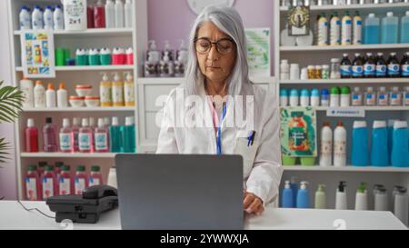 Woman pharmacist with grey hair working on a laptop in a pharmacy store surrounded by various products and shelves indoors Stock Photo