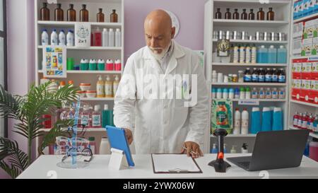 Mature man with a beard working in a pharmacy, wearing a lab coat, checking his tablet while standing behind the counter, surrounded by medicines and Stock Photo