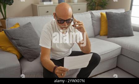 Mature man with grey hair and beard sitting in living room, talking on phone, and reading a document, dressed casually with eyeglasses. Stock Photo