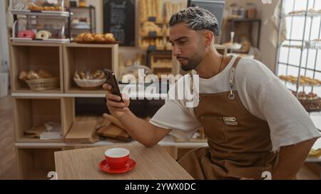 Young man with a beard and apron sitting inside a bakery shop using a smartphone with a cup of coffee on the table and shelves filled with pastries in Stock Photo