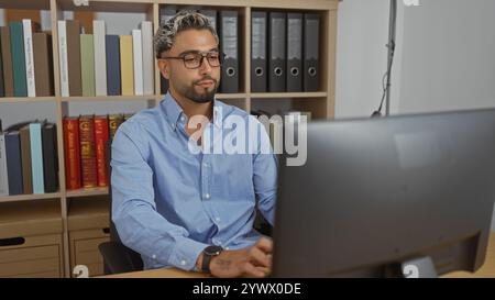 Young, handsome, arab man with a beard working indoors at an office desk, surrounded by documents and shelves filled with colorful books and binders Stock Photo