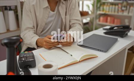 Young hispanic man using smartphone in home decor store, surrounded by notebook, laptop, credit card machine, and bottles Stock Photo