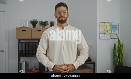 Handsome young hispanic man in a white shirt standing confidently in a modern office workplace with plants and office supplies in the background. Stock Photo