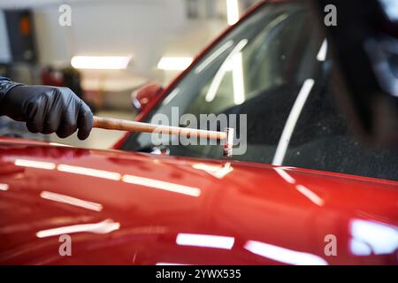 A mechanic repairs a red vehicle with a hammer, highlighting his skill in the busy garage. Stock Photo