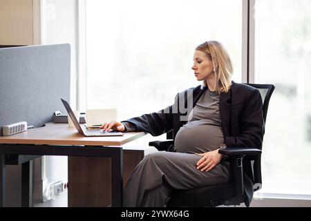Confident pregnant female executive efficiently works on her computer in the office, seated at her desk, dressed in a black blazer, businesswoman over Stock Photo