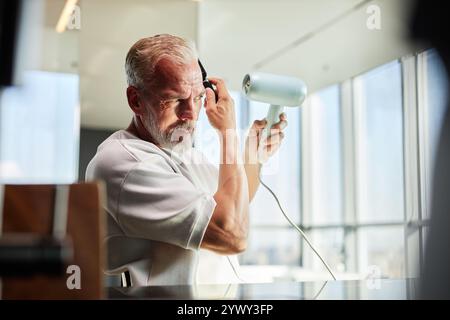 Shot through items on table of mature man concentrated on styling hair with hairdryer after taking refreshing bath as part of daily self care routine at home, copy space Stock Photo