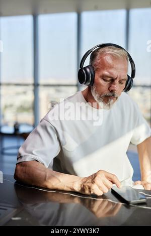 Vertical shot of mature man with neat beard text messaging on phone listening to music in headphones while sitting at table in contemporary apartment, copy space Stock Photo
