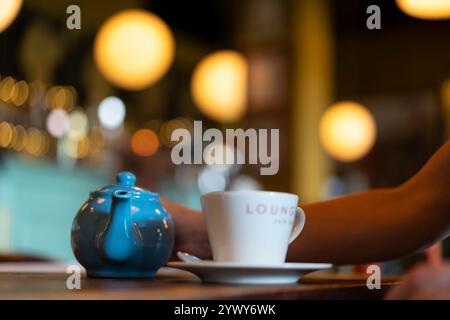 Close up of a woman about to pour tea from a blue, crock tea pot in a restaurant with subtle lighting in the background. Stock Photo