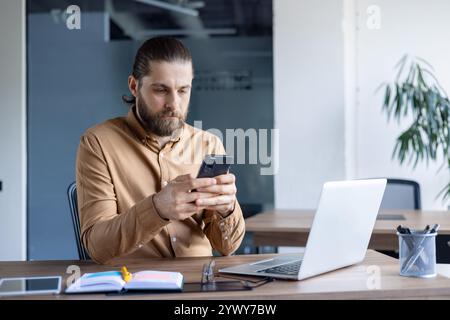A professional dressed in smart casual attire focuses on his smartphone while seated at an office desk, working alongside a laptop and a notepad. Stock Photo