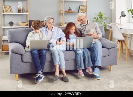 Senior grandparents with grandchildren sitting on sofa using laptops and smartphones together Stock Photo