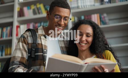African American man Hispanic Latina woman students smile reading book teamwork library knowledge readers club bookshelf university learning studying Stock Photo