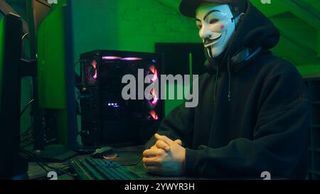 Lviv, Ukraine - June 03, 2024: Masked man anonymous stretching fingers programmer hacking typing computer malware networks web danger scam database Stock Photo