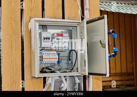 An outdoor metal electrical box with installed circuit breakers and an electricity meter hangs on exterior wall of country house. Stock Photo