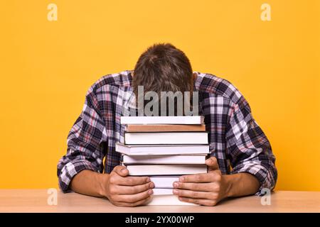 Young student with stack of books having stress before exam at wooden table against yellow background Stock Photo