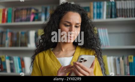 Latin Hispanic Arab woman student mobile phone user smartphone texting sms chatting gadget library university high school college networking smile Stock Photo