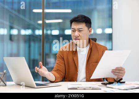 Asian man examining documents with a critical expression while using a laptop in office, reflecting a moment of professional questioning. Stock Photo
