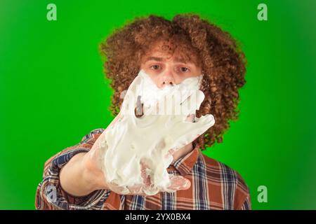 His face partially covered with foam, the curly-haired boy dressed in a neat shirt rubs it gently, standing confidently against the vibrant green Stock Photo