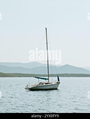 Small sailing boat anchored on calm sea with hills in background during summer. Stock Photo