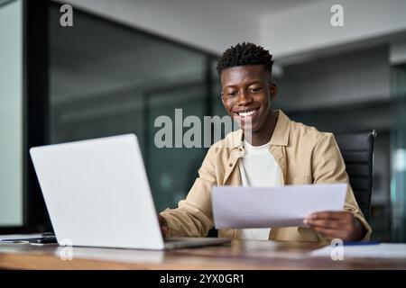 Happy young African business man holding documents using laptop at work. Stock Photo