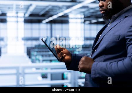 Data researcher using tablet in high tech data center, ensuring cybersecurity and efficient network management. Employee in server hub optimizing data center gear with device Stock Photo