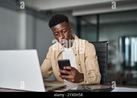 Busy young African business man using smartphone working on laptop in office. Stock Photo