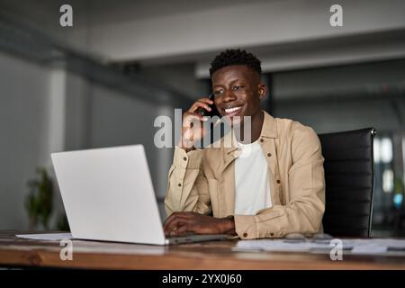 Happy African business man talking on mobile phone working on laptop in office. Stock Photo