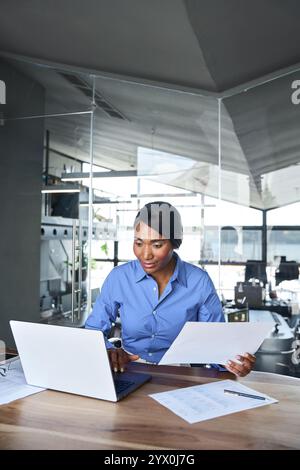 Busy young African American business woman holding papers using laptop. Stock Photo